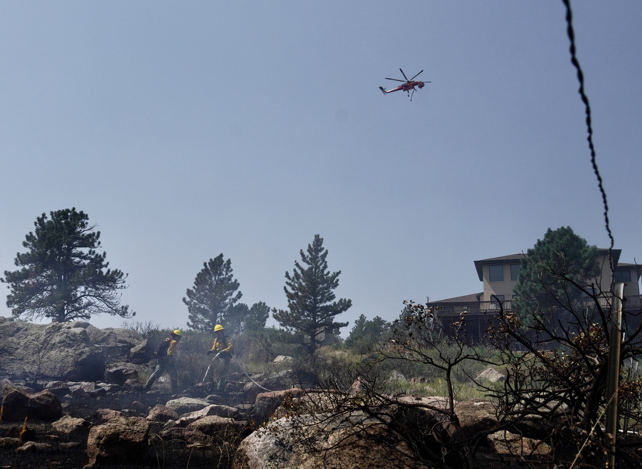 Firefighters douse flames with water near a home being threatened by the Stone Canyon fire as a helicopter flies overhead preparing to dump water on the fire near Lyons on Wednesday, July 31, 2024. (Photo by Zachary Spindler-Krage/The Denver Post)